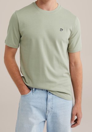 Man draagt een lichtgroen T-shirt met korte mouwen en een klein zwart-wit handlogo, lichtblauwe spijkerbroek, hand in de zak tegen een effen achtergrond.