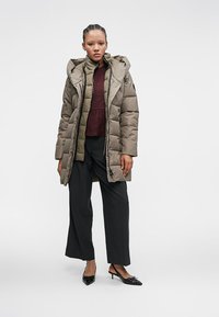 Žena oblečená v dlhom olivovom puffri s kapucňou, bordo topom, čiernych širokých nohaviciach a čiernych lodičkách na podpätku, stojaca na bielom pozadí.