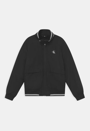 Czarna kurtka z suwakami z kołnierzem stójkowym, z białymi paskami na szyi i u dołu, oraz przednimi kieszeniami. Subtelne logo na piersi.