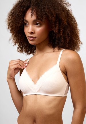 Femme aux cheveux bouclés portant un soutien-gorge rose clair orné de dentelle, touchant la bretelle d'une main, regardant légèrement sur le côté.