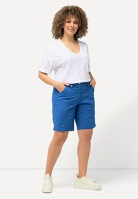 Kvinna med lockigt hår som bär en vit t-shirt med V-ringning, blå knälånga shorts, vita sneakers, står med händerna i fickorna mot en vit bakgrund.