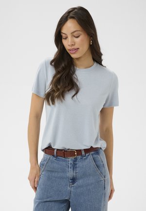 Pikkade tumejate juustega naine, kes kannab helehalli lühikeste varrukatega särki, siniseid teksaseid ja pruunist vööd, vaadates alla ja veidi kõrvale.