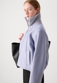 Šviesiai violetinė fliso striukė su aukštu apykakle, užtrauktuku ir šoninėmis kišenėmis. Modelis laiko juodą rankinę. Lygus paviršius, laisvalaikio stilius.