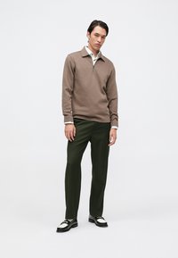 Hombre vistiendo un suéter de cremallera de manga larga marrón, con una camisa blanca debajo, pantalones oscuros verdes y mocasines en blanco y negro, frente a un fondo liso.