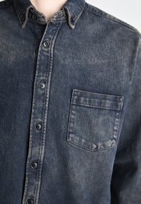 Camicia di jeans di colore blu scuro con texture sbiadita, colletto a dischiudere, taschino frontale e bottoni neri. Presenta un design informale con cuciture visibili.