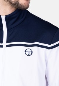 Marineblauwe en witte ritsjack met hoge kraag, textuurstof en horizontale strepen met een rond logo op de borst.