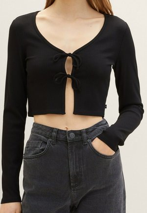 Kobieta ubrana w czarny, długi krótki top z dwoma wiązaniami z przodu, zestawiony z wyblakłymi, czarnymi, wysokim stanem jeansami, z ręką w kieszeni.