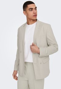 Blazer gris claro de doble botonadura hecho de una tela suave, con solapas con muescas, hombreras y un bolsillo en el pecho. Llevado sobre una camisa blanca.