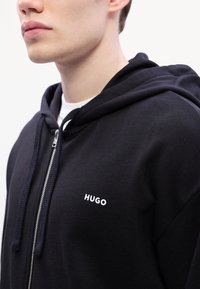 Černá mikina na zip z bavlny, s kapucí se stahovací šňůrkou a bílým logem "HUGO" na levé straně hrudi. Hladká textura, minimalistický design.