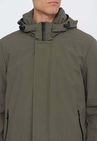 Giacca verde oliva con collo alto, chiusura con zip e cappuccio regolabile. Tessuto liscio e resistente all'acqua con tasche laterali con zip.