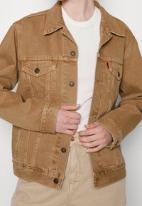 Chaqueta de mezclilla marrón con cierre de botones en el frontal, cuello puntiagudo y bolsillos en el pecho. Presenta una textura en la tela y un diseño sencillo. Usada sobre una camiseta clara.