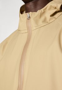 Hellbeige wasserdichte Jacke mit hohem Kragen, Reißverschluss vorne und eingeprägtem Logo. Merkmale sind der glatte, strapazierfähige Stoff und das minimalistische Design.
