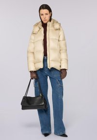 Giacca a vento beige chiaro, con cappuccio e design trapuntato. Abbinata a pantaloni cargo blu denim larghi e guanti neri. Tieni una borsa nera.