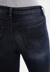 Acercamiento a unos jeans de mezclilla azul oscuro en una persona que lleva una camiseta gris claro, mostrando los bolsillos traseros y la zona de la cintura.