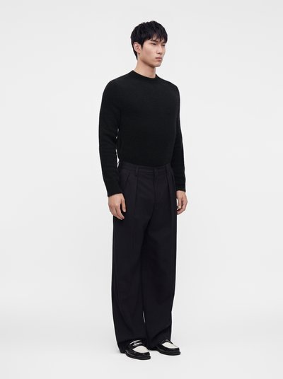Man standing wearing a black sweater, wide black trousers, and black-and-white loafers against a plain white background.