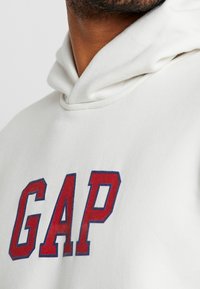 Sudadera blanca con capucha que presenta un logo rojo "GAP" en letra negrita con contornos azules. El tejido parece suave y con textura.