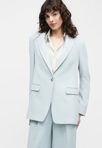 Γυναίκα με σγουρά σκούρα μαλλιά που φοράει ένα ανοιχτό μπλε oversized σακάκι και ταιριαστό παντελόνι με ευρύ πόδι πάνω από λευκό πουκάμισο.