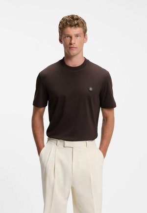 Young man wearing a dark brown short-sleeve shirt tucked into high-waisted beige pleated trousers with hands in pockets.