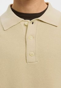 Gros plan d'un polo en tricot beige avec trois boutons, porté par-dessus un t-shirt col rond marron foncé.
