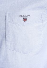Jasnoniebieska, teksturowana koszula z kieszenią na piersi; na kieszeni haftowane logo "GANT" oraz tarcza w kolorach czerwonym, białym i niebieskim.