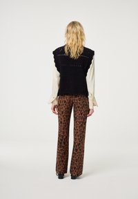 Persoon met blond haar, gekleed in een zwarte mouwloze gebreide spencer, een crème lange mouwen shirt en bruine broek met luipaardprint, staand tegen een witte achtergrond.