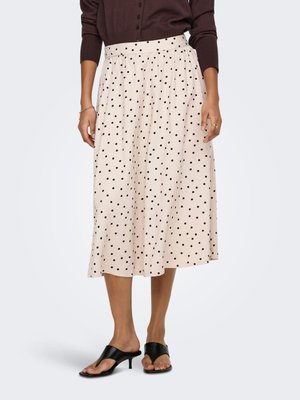 Donna che indossa una gonna midi beige con pois neri, top marrone a maniche lunghe, sandali neri con tacco e gioielli dorati sulle mani.