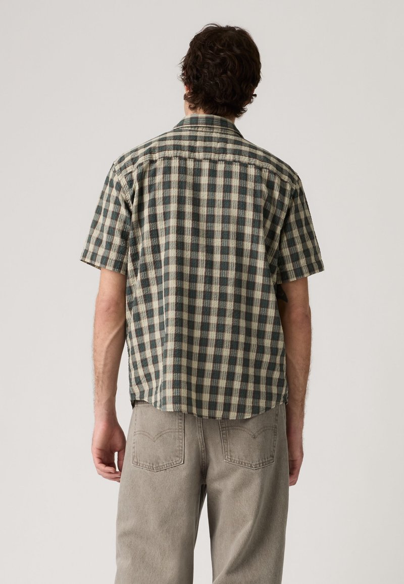 Hombre con camisa de cuadros verde y beige de manga corta y vaqueros gris claro, de pie de espaldas contra un fondo liso.