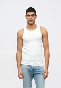 Homem com cabelo curto e preto, vestindo uma t-shirt branca justa e jeans azul-claro, de pé contra um fundo claro e simples.