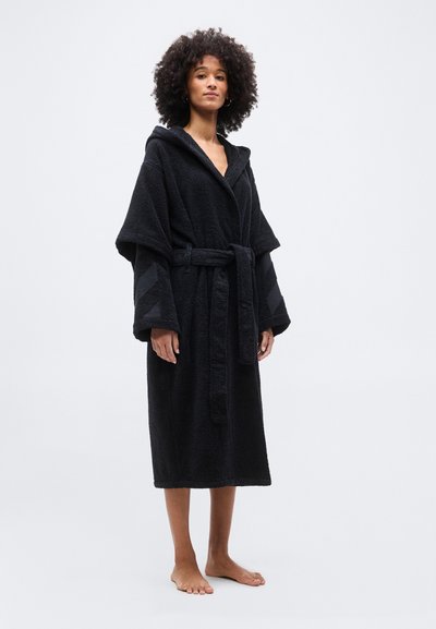 Γυναίκα με σγουρά μαλλιά φοράει μαύρο ρόμπα με κουκούλα, φαρδιά μανίκια και δεμένη ζώνη, στέκεται ξυπόλυτη σε απλό φόντο.