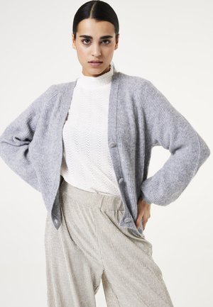 Gray knitted cardigan with a relaxed shape and long sleeves. Features front buttons and a textured white mock neck sweater beneath. Silver striped pants.