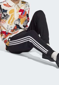 Person som sitter och bär en färgglad t-shirt med grafiskt mönster, svarta Adidas träningsoverallbyxor med vita ränder, vita strumpor och svarta sneakers.