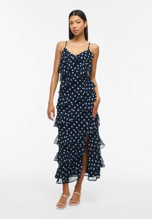 Femme portant une robe maxi sans manches bleu marine à pois, avec des volants et une fente avant, associée à des talons ouverts blancs.