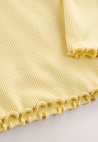 Žuto rebrasto tkivo s valovitim, naboranim rubom. Glatka tekstura s okomitim linijama, presavijeno na jednom kutu.