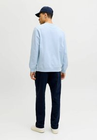 Šviesiai mėlynas džemperis su gofruotais rankogaliais ir apačia, derinamas su tamsiai mėlynomis kelnėmis. Modelis dėvi tamsią kepurę ir kreminius batus, demonstruodamas laisvą stilių.