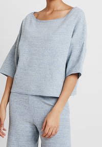 Helles blaues, strukturiertes Oberteil mit weitem Ausschnitt und Dreiviertelärmeln, das einen lockeren Sitz und ein weiches, gestricktes Material aufweist.