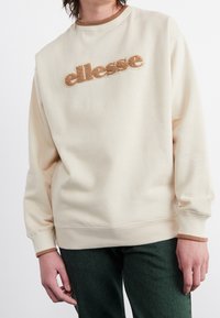 Krämfärgad sweatshirt med ribbad halsringning och manschetter, med en upphöjd brun logotyp över bröstet. Mjukt tyg med en avslappnad passform.