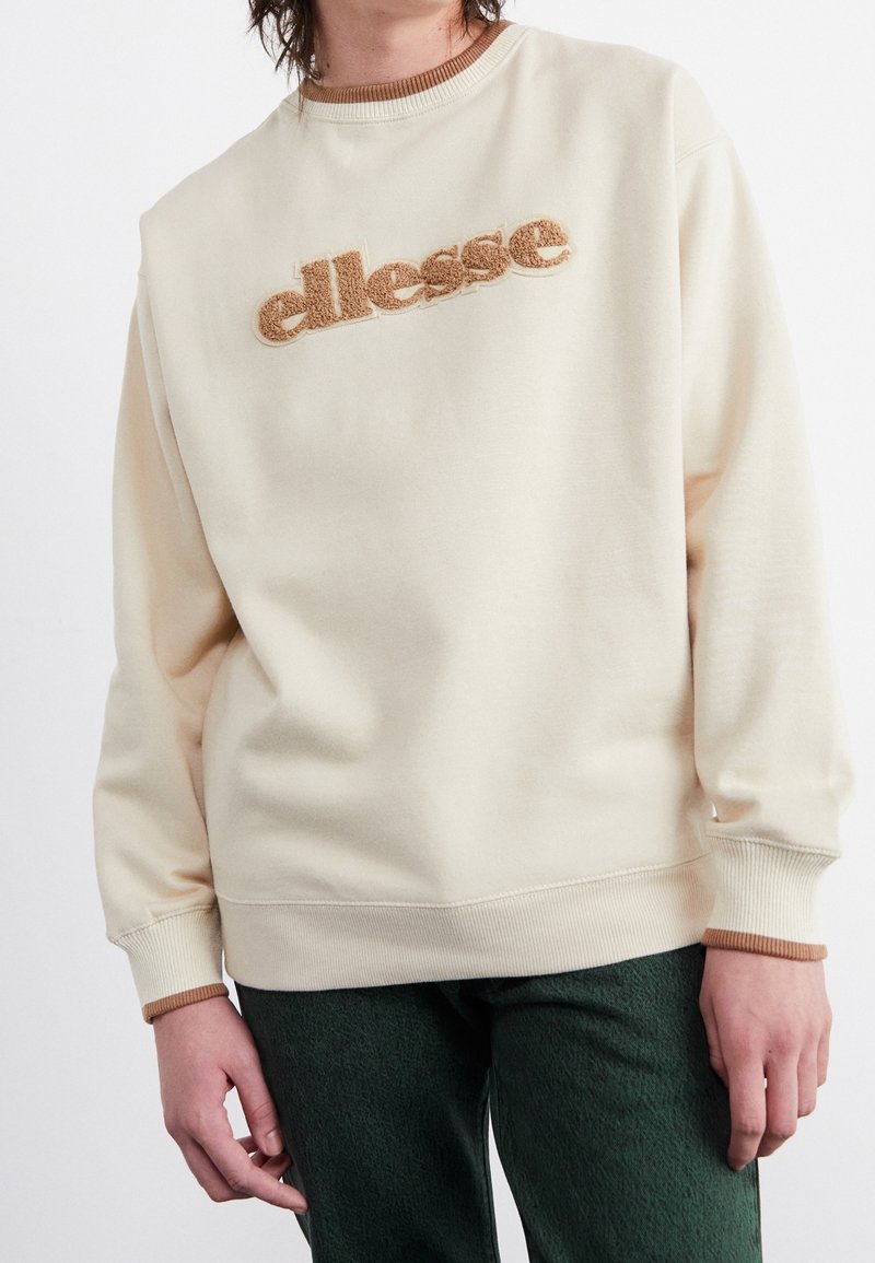 Krämfärgad sweatshirt med ribbad halsringning och manschetter, med en upphöjd brun logotyp över bröstet. Mjukt tyg med en avslappnad passform.