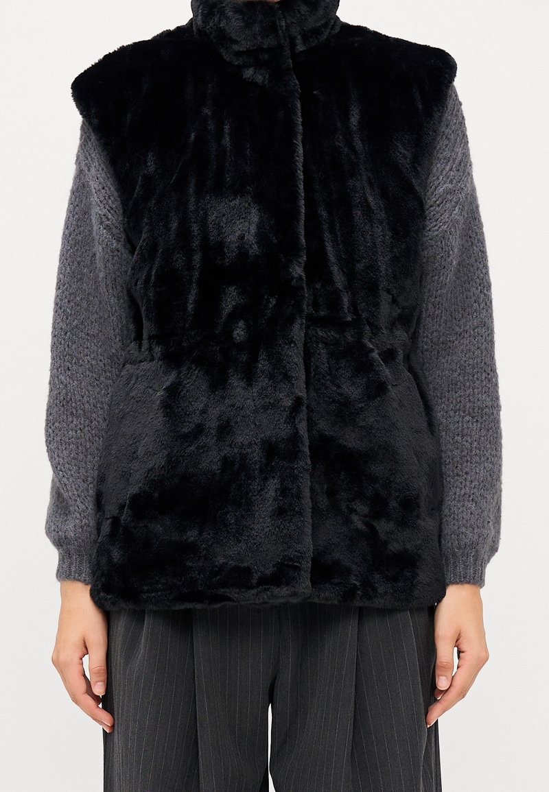 Personne portant une veste noire en fausse fourrure sur un pull gris tricoté avec un pantalon gris foncé à fines rayures, debout devant un fond blanc.