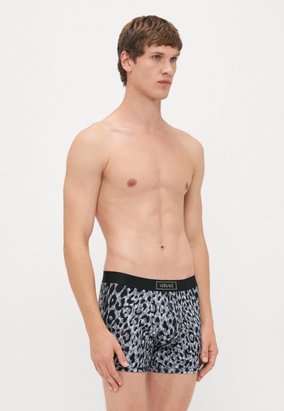 Skjorteløs mannemodell iført grå og svart leo-print Versace boxershorts med svart midjebånd, stående mot en enkel hvit bakgrunn.