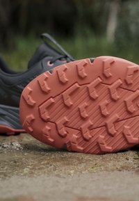 Chaussure de trail running noire avec une semelle robuste orange vif, inclinée sur un sol en terre à l'extérieur.