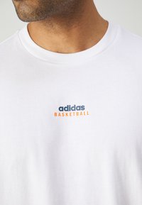 Hvit bomull T-skjorte med ribbet rund hals. Har "adidas"-logoen i blått og "BASKETBALL" i oransje på venstre brystområde.