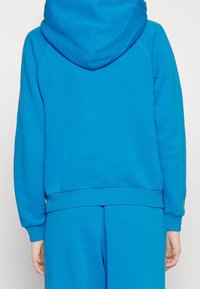 Blå munkjacka gjord av mjukt material, med dragsko i huvan, ribbade muddar och nederkant, samt en avslappnad passform. Vy från baksidan.