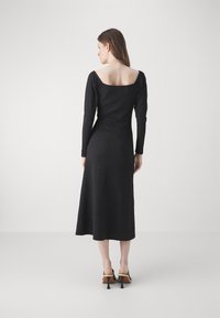Black long-sleeve dress with a square neckline, fitted bodice, and flared skirt. Textured fabric and mid-length hem. Worn with high-heeled shoes.