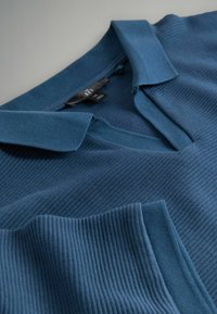 Blå ribstrikket sweater med V-hals og krave, vist foldet med synlig sort størrelsesetiket.