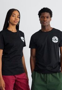 Melni īsām piedurknēm t-krekli ar baltu "ALPHA INDUSTRIES" grafiku priekšā, apvienoti ar sarkanām un zaļām biksēm. Vienkārša balta fona.
