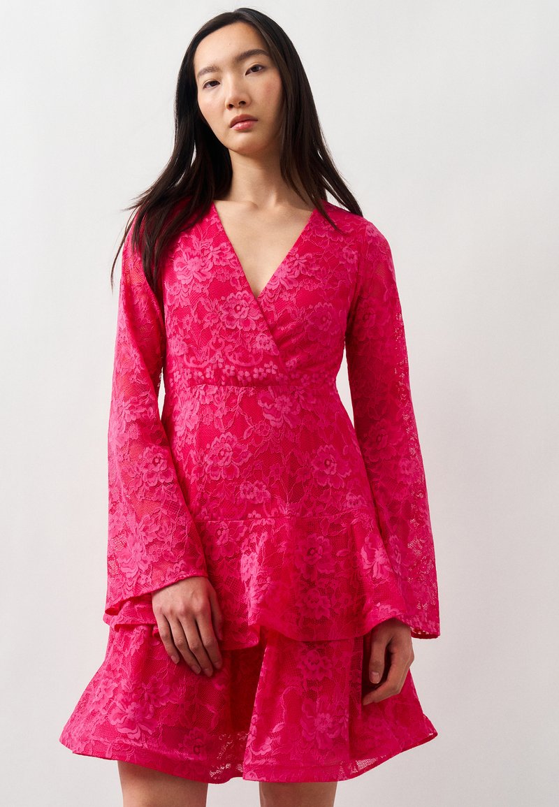 Νεαρή γυναίκα φοράει ένα φωτεινά ροζ λουλουδάτο φόρεμα με δαντέλα τύπου wrap, με μακριά μανίκια καμπάνα και φούστα με βολάν, σε απλό λευκό φόντο.