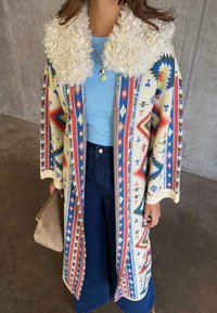 Άτομο που φοράει παλτό με μοτίβο και μεγάλο λευκό χνουδωτό γιακά, ανοιχτόγαλου ζακέτα, σκούρα μπλε τζιν και κρατάει μπεζ τσάντα χειρός.