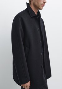 Cappotto di lana nera con colletto, chiusura frontale con bottoni e tasche laterali, dal taglio rilassato e con dettagli di cucitura minimi.