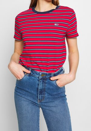Femme portant un t-shirt à manches courtes rayé horizontalement rouge, bleu marine et blanc, rentré dans un jean bleu taille haute, les mains dans les poches.