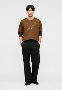 Άνδρας με μαύρα μαλλιά, φορώντας καφέ πουκάμισο με μακριά μανίκια και λευκό σχέδιο και μαύρο παντελόνι, στέκεται με τα χέρια στις τσέπες σε απλό φόντο.